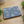 Rectangular tray with a blue and white yukiwa pattern featuring delicate floral motifs inside stylized snow rings, set against fine vertical stripes. The tray rests on a light wood surface surrounded by green foliage.