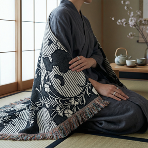 Woven blanket with a dark blue and off-white katagami-inspired pattern featuring stylized chrysanthemums, leaves, and stripes. The blanket has a multicolored fringe and is draped over a person sitting on a tatami mat.