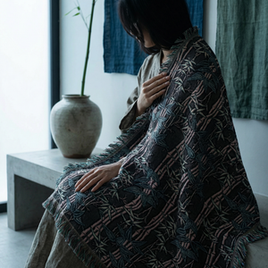 Person wrapped in a patterned blanket sitting in a room with a vase and curtains.