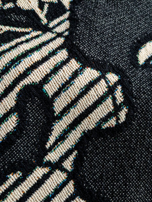 Close-up of a woven blanket featuring a traditional Japanese katagami design in black and off-white. The pattern includes stylized botanical motifs such as slender branches with small leaves or plum blossoms.