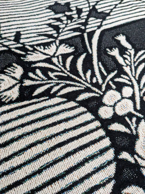 Close-up of a woven blanket featuring a traditional Japanese katagami design in black and off-white. The pattern includes stylized botanical motifs such as slender branches with small leaves or plum blossoms.
