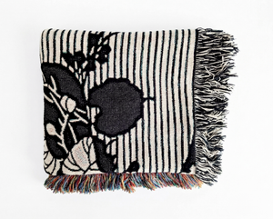 A folded woven blanket featuring a large-scale black botanical pattern of broad, rounded leaves and delicate stems, set against a background of vertical dark blue and off-white stripes.