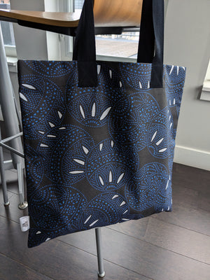 Black tote bag with a dynamic circular pattern in blue and white, featuring dotted and curved elements. Made from organic cotton canvas, ideal for stylish daily errands.