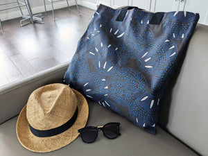 A black tote bag with a blue and white circular pattern featuring dotted and curved elements, styled on a gray sofa next to a straw hat with a black band and black sunglasses.