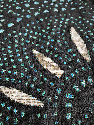 Woven blanket with a black background featuring scattered, irregular clusters of blue, green, and white threads.