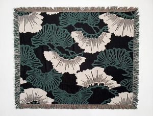 Woven blanket featuring a repeating Japanese katagami pattern of stylized pine (matsu) motifs. The design includes fan-like clusters of teal green pine needles and larger, abstract off-white shapes.