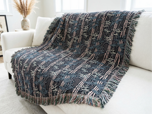 Woven blanket featuring a Japanese bamboo (take) motif with pale pink stalks and leaves in pale sage and muted teal, set against a dark grey background. The blanket is draped over a white sofa and has multi-colored fringe.