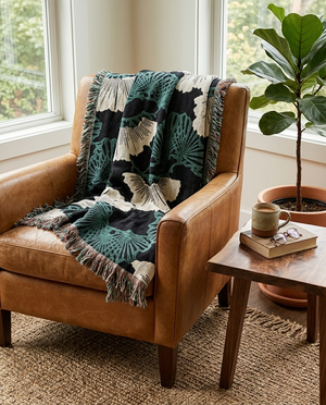 Brown leather armchair with a patterned green blanket, a wooden side table with a mug and book, and a potted plant in the background.