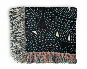 Folded woven blanket with a black ground featuring scattered irregular light blue dots and abstract off-white triangular shapes. The left fringe is black and cream, and the bottom fringe is multi-colored.
